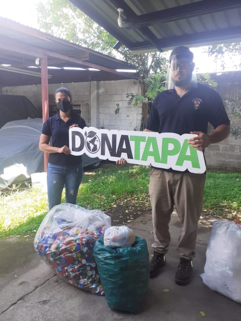 Hombre y mujer con máscara COVID sosteniendo rótulo Donatapa a la par de bolsas llenas de tapas